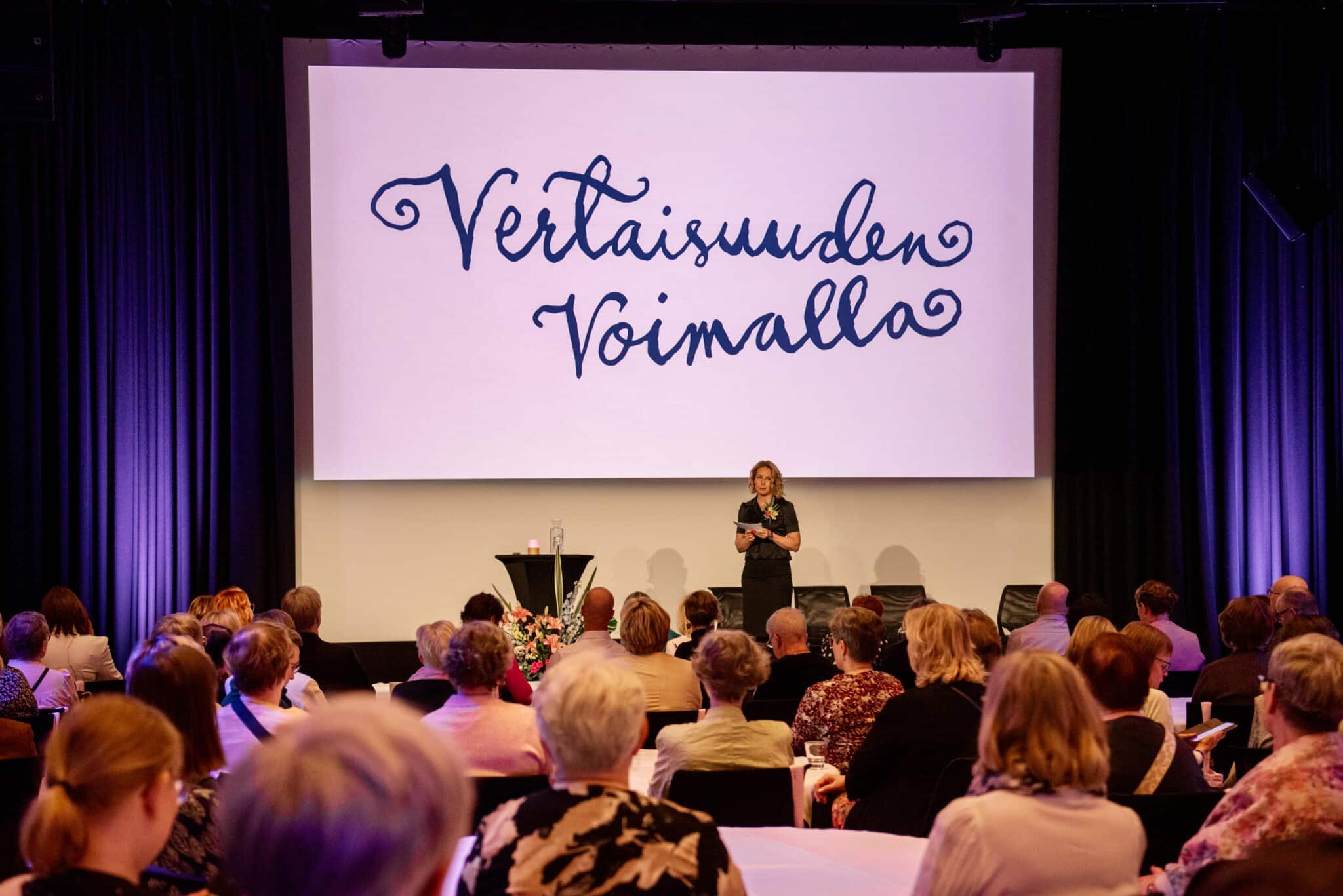 Rintasyöpäyhdistys - Europa Donna Finland ry:n 20-vuotisjuhlassa sali täynnä ihmisiä istumassa pöytien ääressä ja valkokankaalle heijastettuna teksti "Vertaisuuden voimalla"