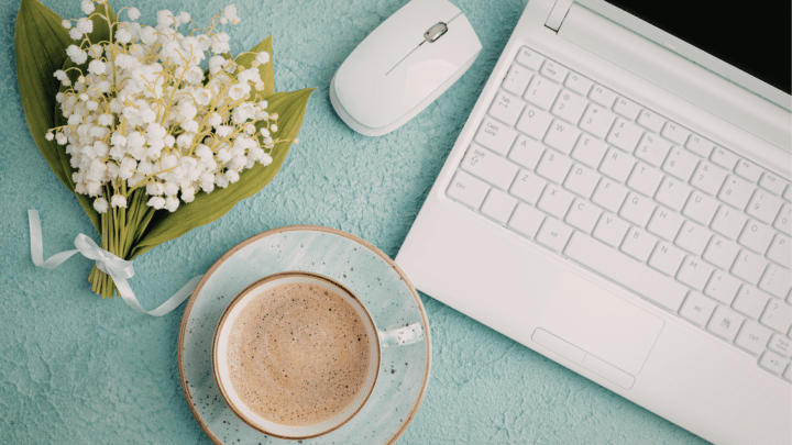 Vaaleansinisellä pöydällä kimppu kieloja, kupillinen kahvia ja kannettava tietokone sekä irrallinen tietokoneen hiiri.